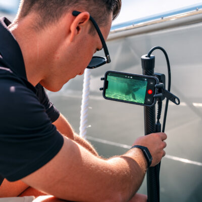 Live underwater view from the Seavu Seeker Pole Kit shown on a phone mounted to the carbon-fibre pole — a technician watches and records real-time footage during a hull inspection