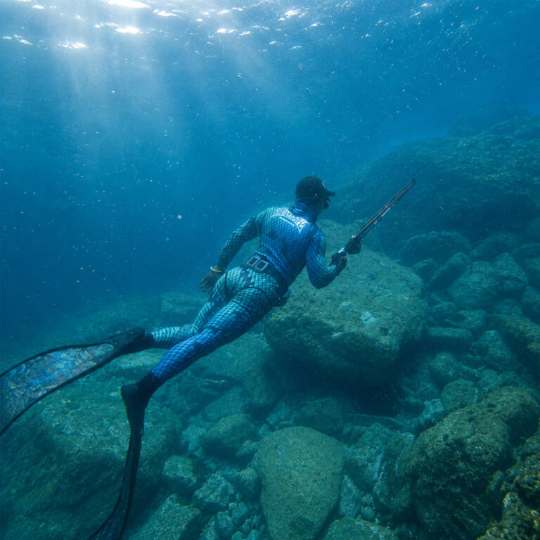 Spearfisher diving over a rocky reef with a speargun, lit from the surface
