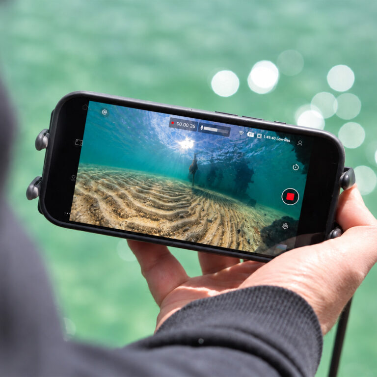 Phone in a Seavu phone mount showing live 360 underwater footage of sand ripples and jetty pylons from the Seavu Orbit 360